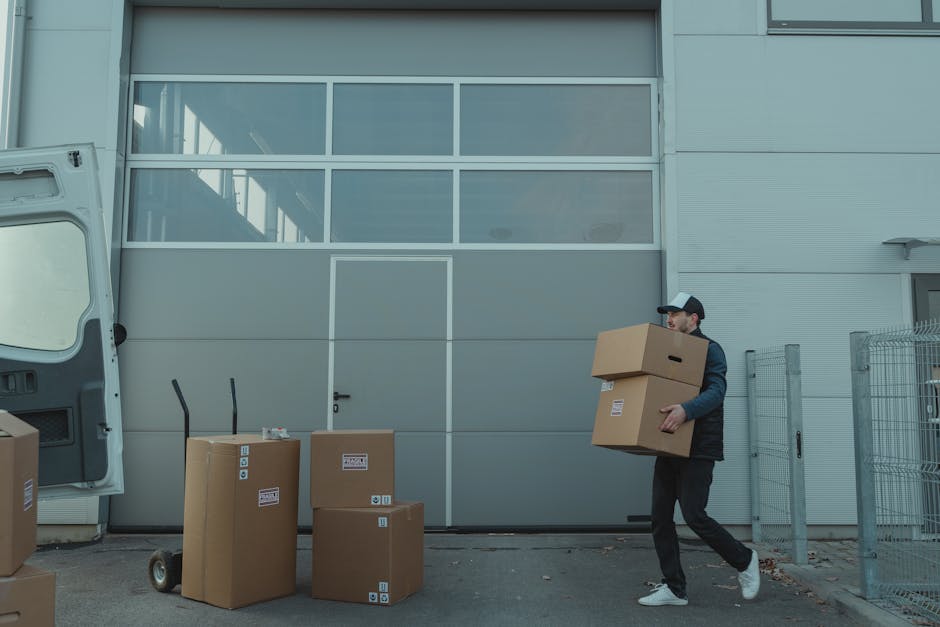 A man wearing a black cap, dark jacket, and black trousers is carrying a cardboard box while walking away from a large industrial building with gray paneling and glass windows. Several other cardboard boxes, some sealed with packing tape and others with labels, are positioned on the pavement nearby, along with a small hand truck. The open rear doors of a white van are visible on the left side of the image, indicating the loading or unloading activity associated with house removals or furniture transport. The scene suggests an organized home relocation process, with the man preparing to load the boxes into the vehicle as part of a professional moving service. The environment is well-lit, with clear details of the boxes, equipment, and the exterior of the building, supporting the context of packing and moving logistics for a house removal, consistent with the services offered by Beckenham Man and Van.