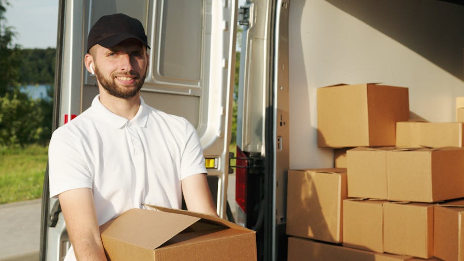 A man with a beard, wearing a black cap and a white polo shirt, is standing outside next to an open van used for home relocation. He is holding a cardboard box in his hands, which appears ready for loading or unloading. Inside the van, there are neatly stacked cardboard boxes of various sizes, some wrapped in plastic or packing materials, ready for transport. The background shows a natural outdoor setting with greenery and a clear sky, indicating bright daylight. The van's interior is partially visible, showing a clean and organized space for furniture transport and packing materials. This scene captures the loading process typical of window-based house removals, with the man preparing to assist in furniture and box transfer, showcasing the logistical aspects of professional removals. The setting emphasizes the moving process, packing, and loading within a professional service environment, relevant to beckenhammanandvan.com’s house removal services for flats and homes.