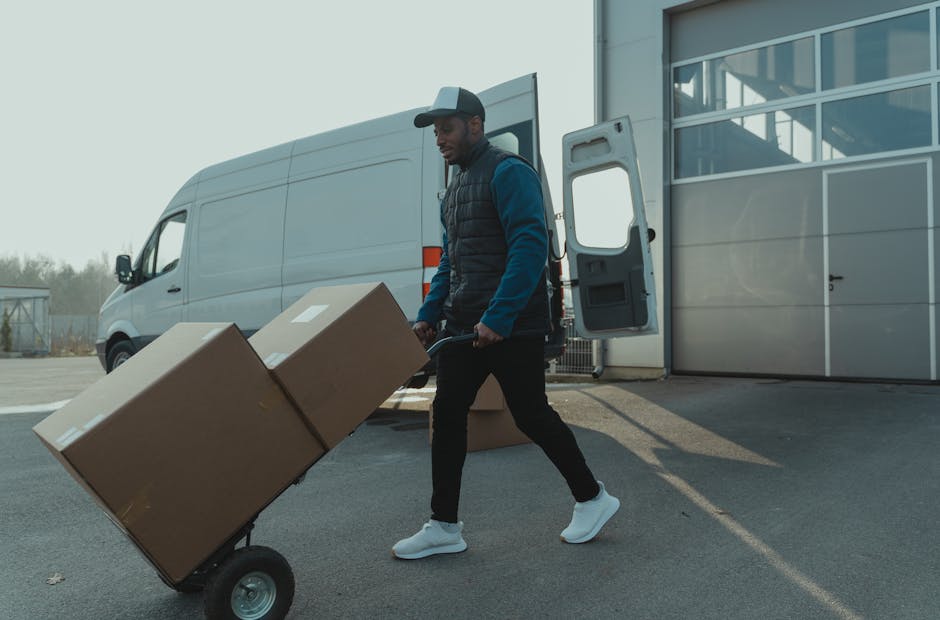 A man wearing a black cap, dark jacket, and black trousers is carrying a cardboard box while walking away from a large industrial building with gray paneling and glass windows. Several other cardboard boxes, some sealed with packing tape and others with labels, are positioned on the pavement nearby, along with a small hand truck. The open rear doors of a white van are visible on the left side of the image, indicating the loading or unloading activity associated with house removals or furniture transport. The scene suggests an organized home relocation process, with the man preparing to load the boxes into the vehicle as part of a professional moving service. The environment is well-lit, with clear details of the boxes, equipment, and the exterior of the building, supporting the context of packing and moving logistics for a house removal, consistent with the services offered by Beckenham Man and Van.
