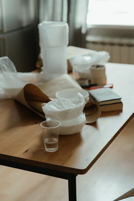 A wooden table inside a home contains various packing and moving materials, including several large sheets of bubble wrap, tightly rolled and partially unrolled, along with cardboard packing tubes and rolls of adhesive tape. There are also a couple of books stacked beside the packaging supplies, and a small clear plastic cup or glass placed on the table. The background features a window with natural light illuminating the scene and a radiator underneath, indicating a domestic environment prepared for packing and home relocation by [COMPANY_NAME], such as Beckenham Man and Van, during a furniture transport or moving process for the Kelsey Park house removals guide for fragile items.