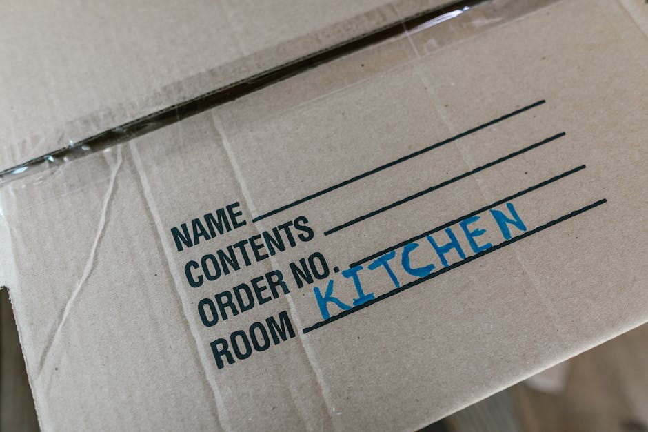 Close-up view of a brown cardboard moving box with a white label on the top left corner, which has fields for name, contents, order number, and room; the room field is handwritten in blue ink as 'KITCHEN'. The box appears to be part of a house removal process, with packing materials possibly visible around it. The box is situated indoors near a doorway or entry point, and in the background, there may be hints of additional packed boxes or household items ready for transport. This image illustrates the packing stage typical of home relocation services provided by Beckenham Man and Van, including organization of fragile or important items for a furniture transport and loading process.